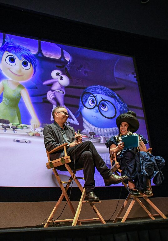 Two panelists sit in directors chairs on stage in conversation. Behind them, a large screen with characters from Pixar's INSIDE OUT is projected.