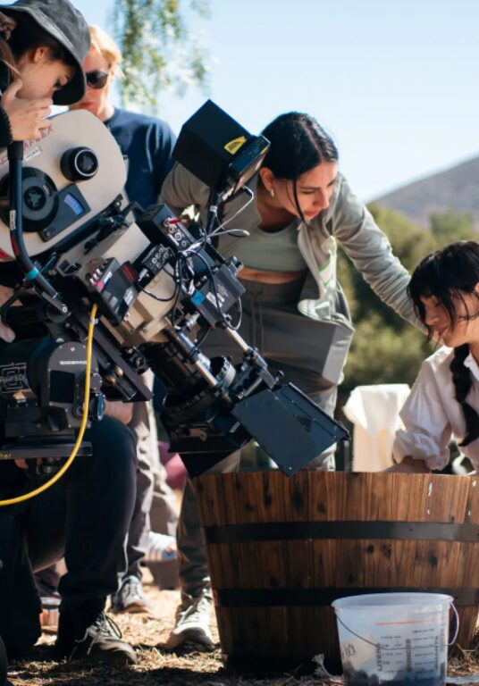 A small film crew gathers around a large cinema camera on a wheeled rig, filming a scene outdoors. A woman in a white blouse and long skirt kneels beside a wooden tub, smiling as a crew member adjusts her position. Trees and hills are visible in the sunny background, suggesting a rural setting.