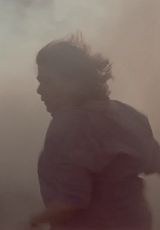 A hazy, motion-blurred image of a person in profile moving through a dusty or smoky environment, their hair blown back and a short-sleeved shirt visible, creating a sense of urgency or movement.