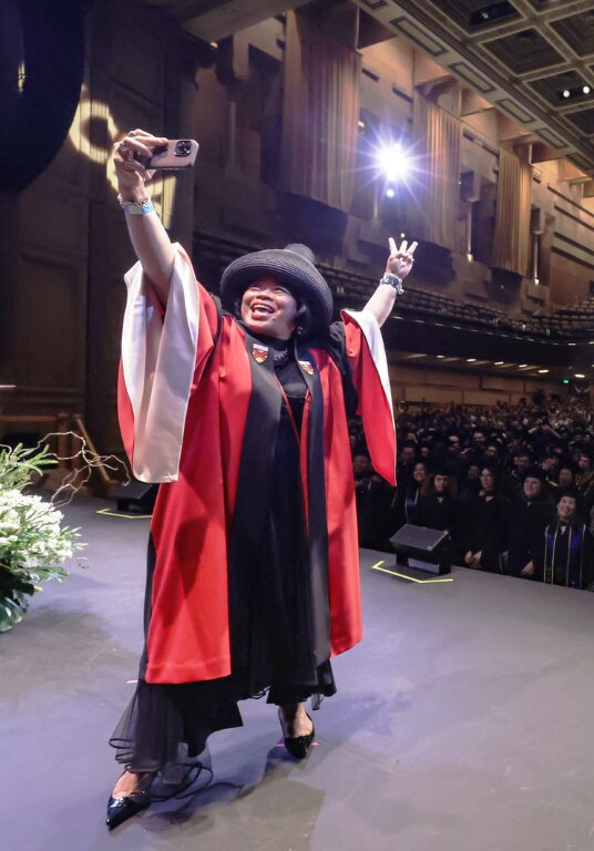 A woman in red graduation robes stands on a graduation stage taking a selfie with the audience behind her