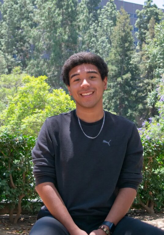 Boy in black shirt and chain necklace sits outside in daylight on a bench in front of a lush green background of trees