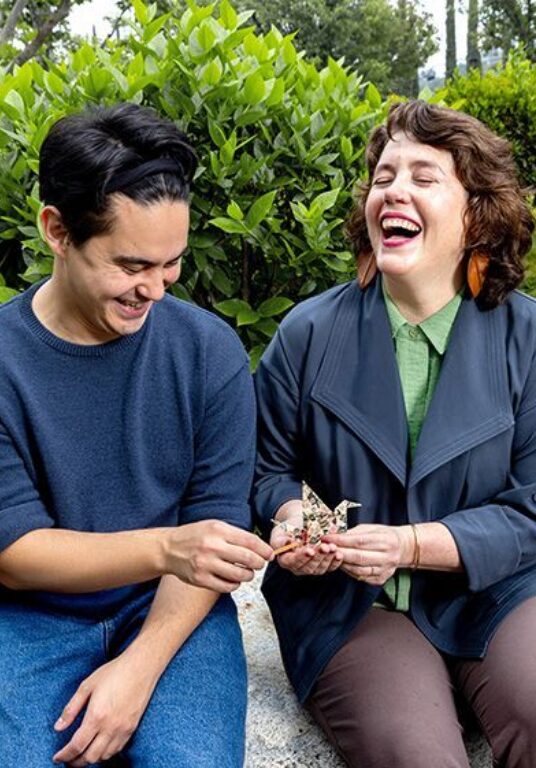 Female teacher and male student sit outside together with a small puppet in hand, laughing