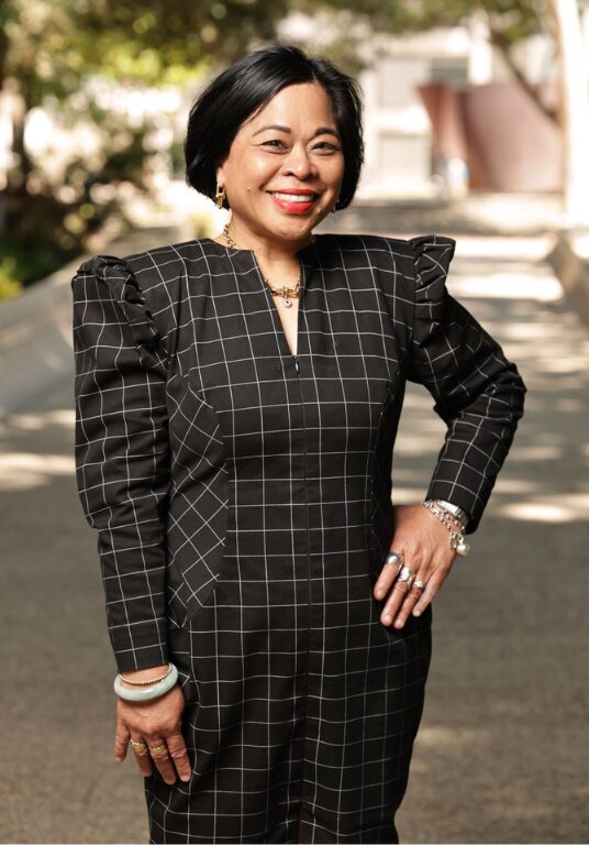 A woman with short black hair in a black and white grid-patterned skirt suit poses with a hand on her hip in the UCLA Sculpture Garden
