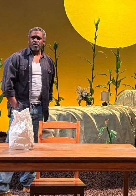 Black man on a yellow-washed stage stands in a dark button up shirt and white tank top with the sun behind him