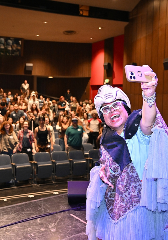 A woman takes a selfie on stage with a crowd of people behind her inside an auditorium