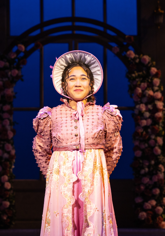 A young girl stands on stage in a pink bonnet and Regency-era dress.