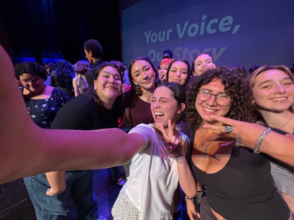 A group of female students pose together for a selfie in front of a background on stage that reads Your Voice, Our Story.