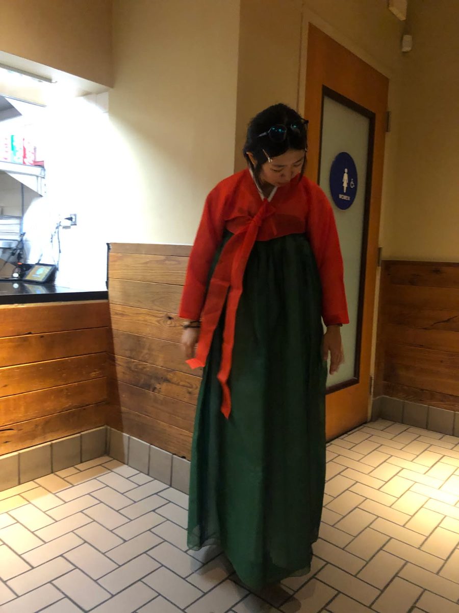 A woman stands in a warm lit restaurant wearing a traditional red and green hanbok from South Korea
