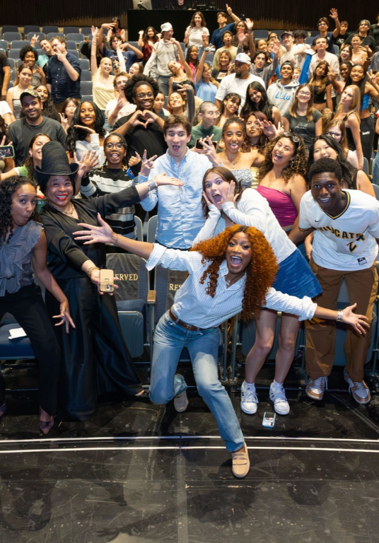 A crowd of students in an auditorium. The crowd has their arms spread wide.
