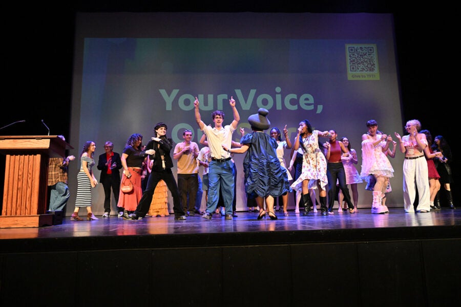 A group of people dance on a stage in front of a backdrop with the words Your Voice, Our Story