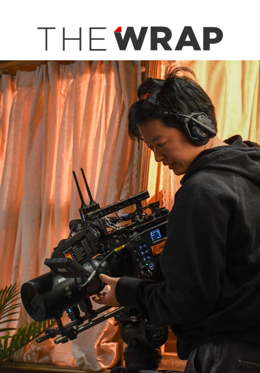 TheWrap logo above a photo of a camera operator wearing headphones and adjusting a professional film camera on set.